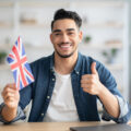 Smiling guy with flag of Great Britain showing thumb up Smiling arab guy with flag of Great Britain showing thumb up, sitting at desk with laptop, middle-eastern young man student learning english for job, education or emmigration to UK, copy space